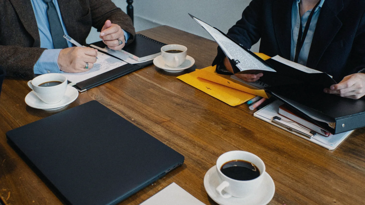 Two people across a wooden table with coffee, one taking notes, the other opening a binder; no faces shown