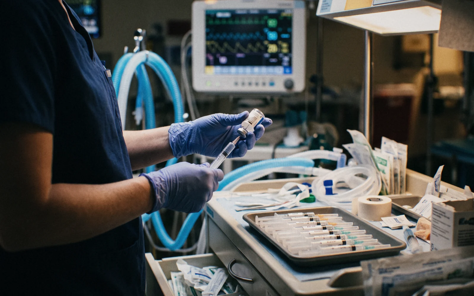 An anesthesia provider drawing up medication at the anesthesia cart, the monitor and breathing circuit behind